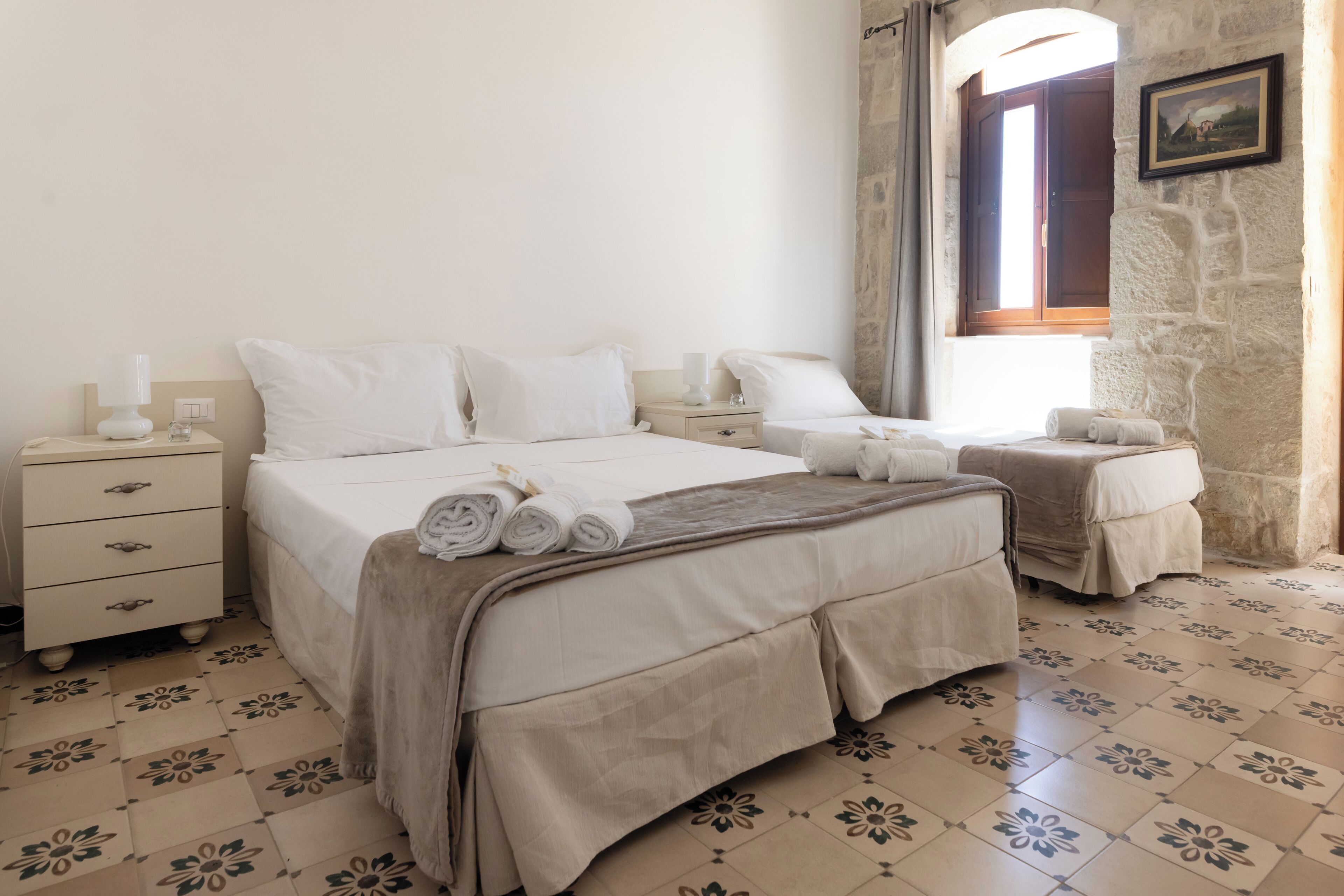 Une chambre avec un grand lit, deux tables de chevet, une commode, une fenêtre avec des rideaux et un mur en pierre.