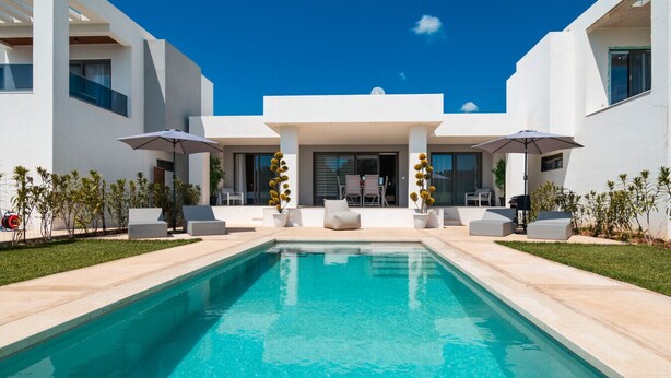 Een modern zwembad met helderblauw water, witte tuinmeubels en een overdekte zitplaats.
