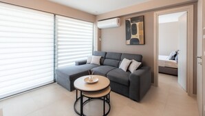Een moderne woonkamer met een grijze bank, een ronde tafel en een groot raam met zonwering.