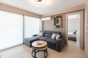 A modern living room with a grey sofa, a round table, and a large window with blinds. - Luna Villas Moon by RentalsPro - Portaria Beach (Nea Moudania)