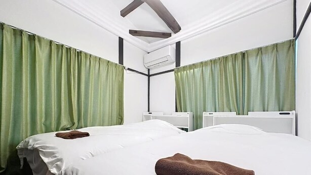 A hotel room with two beds, green curtains, white bedding, and an air conditioning unit. - Q stay Toneri Park (Tokyo)