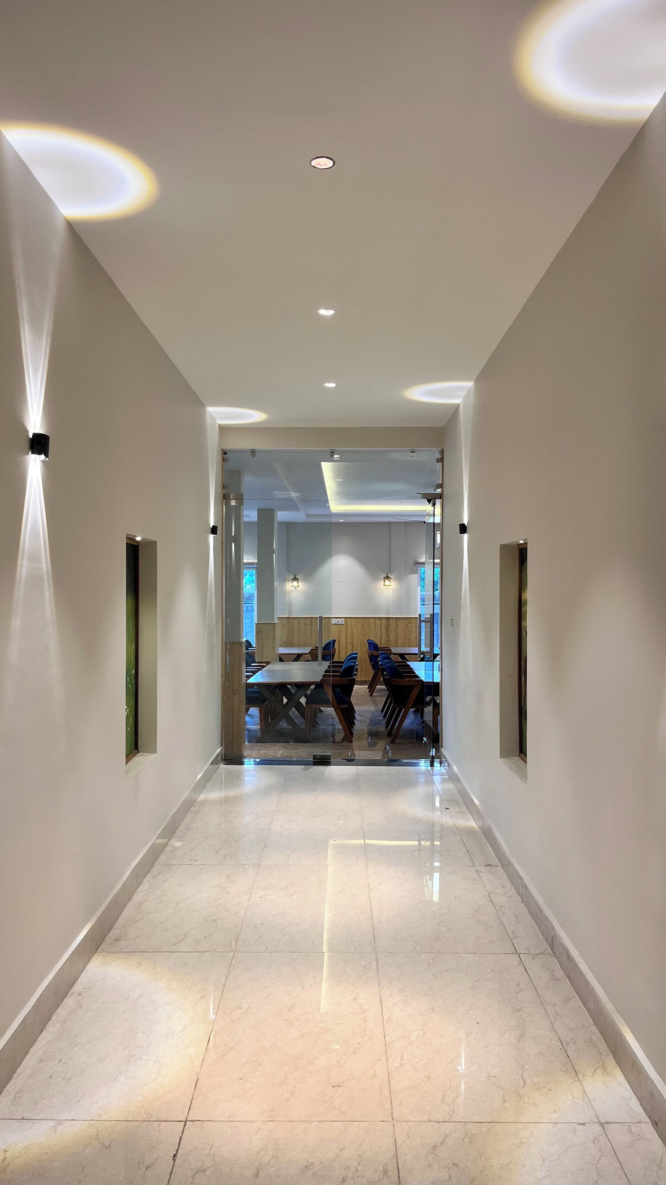 Un pasillo moderno de hotel con suelo de mármol pulido, iluminación empotrada y vista a un comedor con mesas y sillas.
