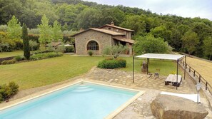Una casa de piedra con piscina, rodeada por un jardín y una ladera boscosa.