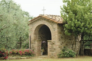 A stone chapel with an arched entrance, a cross on the roof, and a garden with flowers and trees. - Panoramic villa between Umbria and Tuscany (San Leo Bastia)