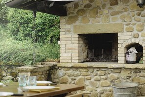 Una chimenea de piedra al aire libre con un estante de ladrillo arriba, una mesa con vasos y un cuaderno, y un cubo de metal.