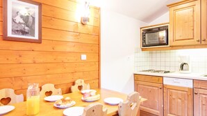 A wooden dining table with chairs, plates, and a glass of juice. There is a framed picture on the wall, wooden cabinets, and a microwave in the kitchen area. - Cosy Apartment in Modane by Ski-lifts (Modane)