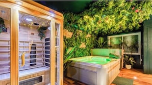 Eine Sauna mit Glastür und Holzinnenraum, eine grüne beheizbare Wanne und eine Wand mit künstlicher Bepflanzung.