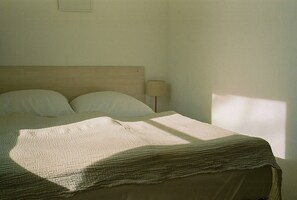 Sänky, jossa on puinen pääty, lamppu ja ikkuna, josta valo osuu sängylle.