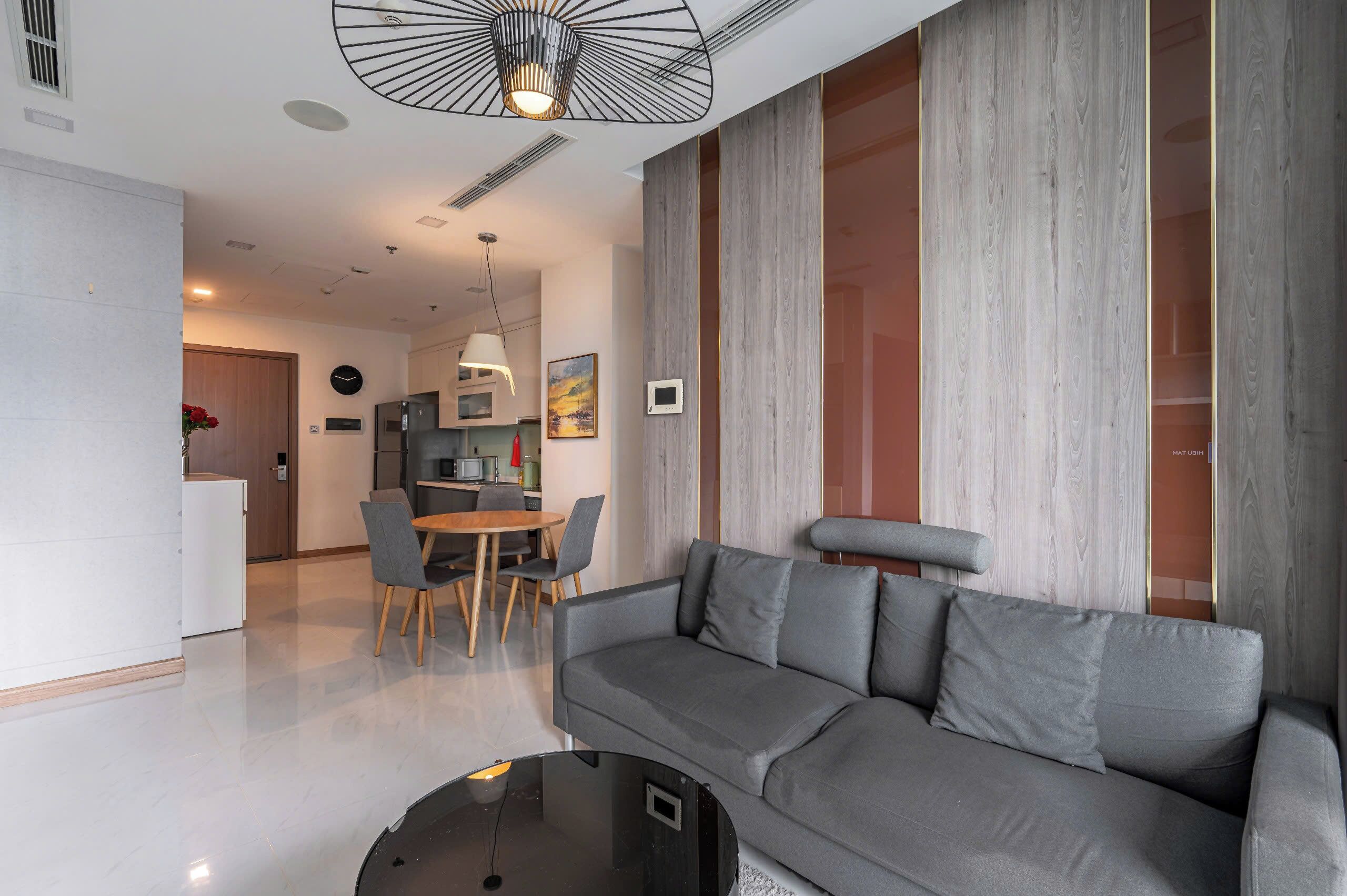 A modern living room with a grey sofa, a dining table with chairs, a kitchen in the background, and a large window with curtains.