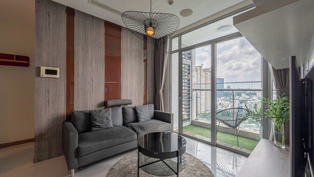 A modern living room with a sectional sofa, a round coffee table, a pendant light, a large window with a view of the city, and a small plant on the windowsill. - The Grand K-Vinhomes - Stunning Views (Ho Chi Minh City)