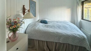 Ett sovrum med en säng, en byrå, en lampa, en vas med blommor och ett fönster med gardiner.