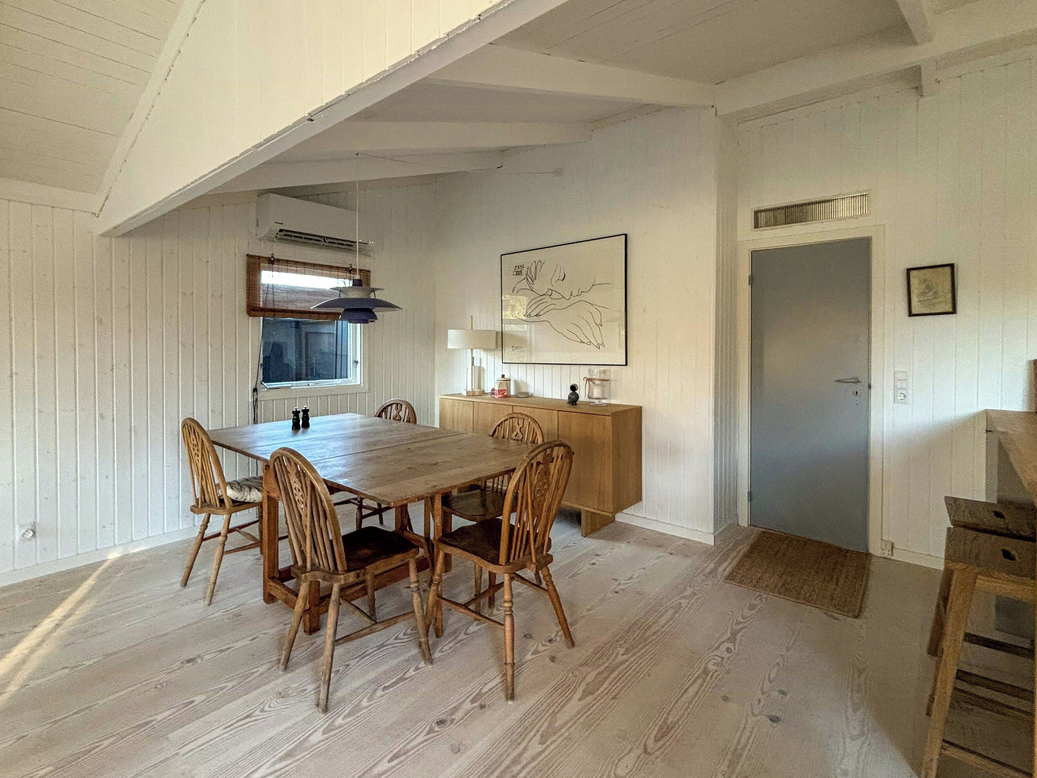 A dining area with a wooden table and chairs, a window, a wall-mounted lamp, and a framed picture.