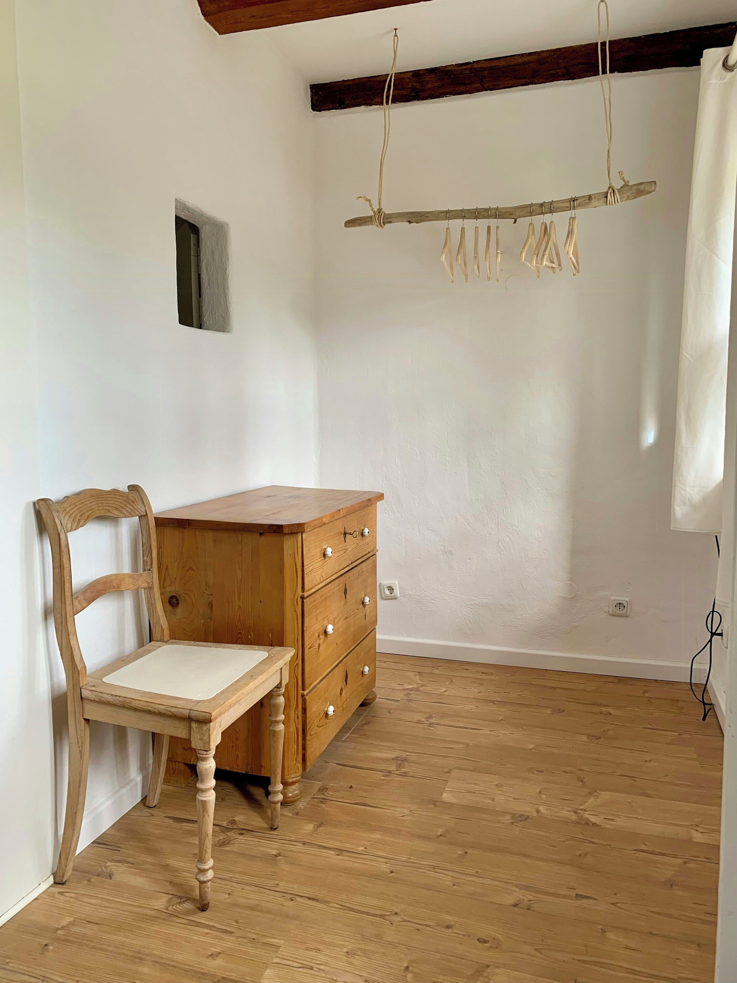 A room with a wooden chair, a wooden dresser, a small window, and a wooden beam ceiling.