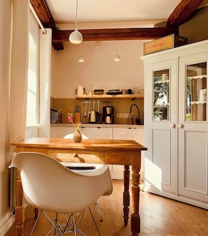 A kitchen with wooden beams, white cabinets, and a dining area with a white chair. - Charming garden apartment in an old thatched house (Steinberg)