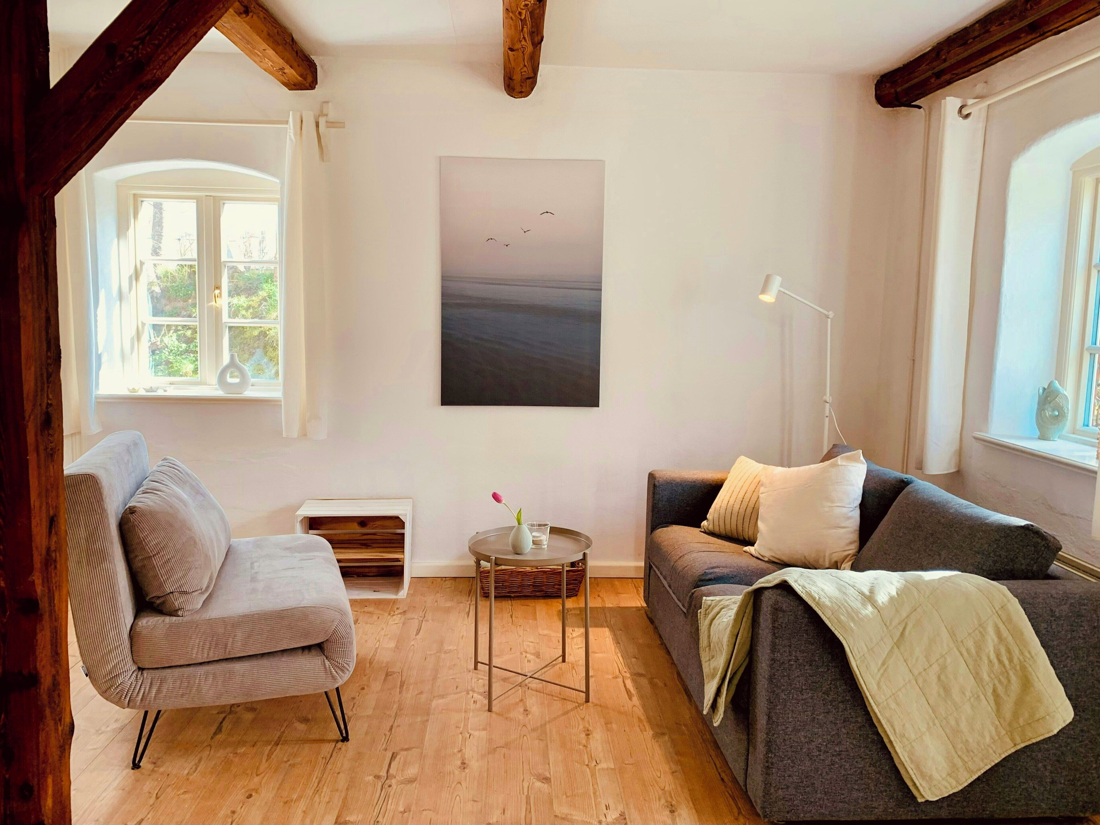 A room with a wooden ceiling, a painting of birds, a sofa, a chair, a small table, and a lamp.