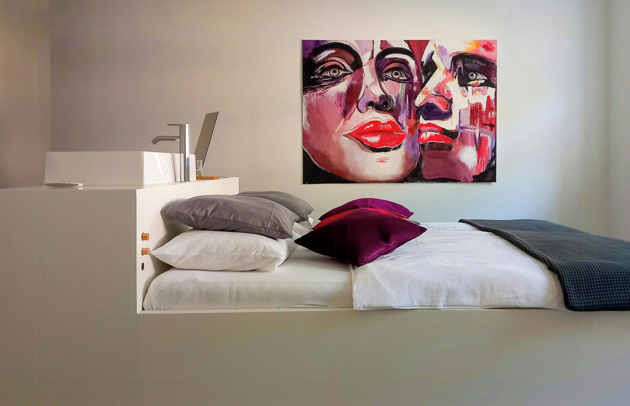 Un dormitorio moderno con cama, almohadas, mesita de noche, espejo y un cuadro colgado en la pared.