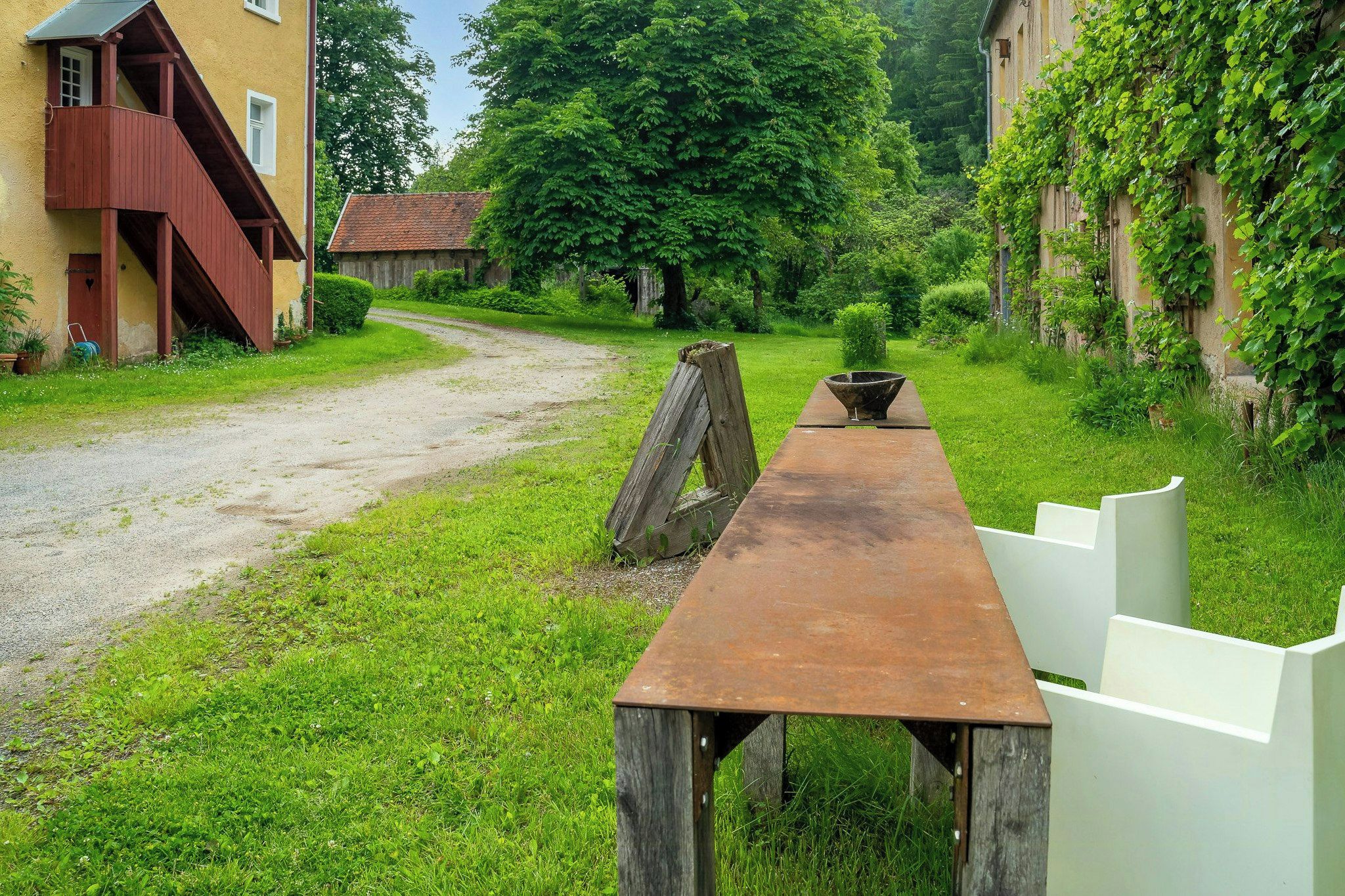 Eine ländliche Außenanlage mit einem Holztisch und Bänken, umgeben von Grünpflanzen und einem Kiesweg.