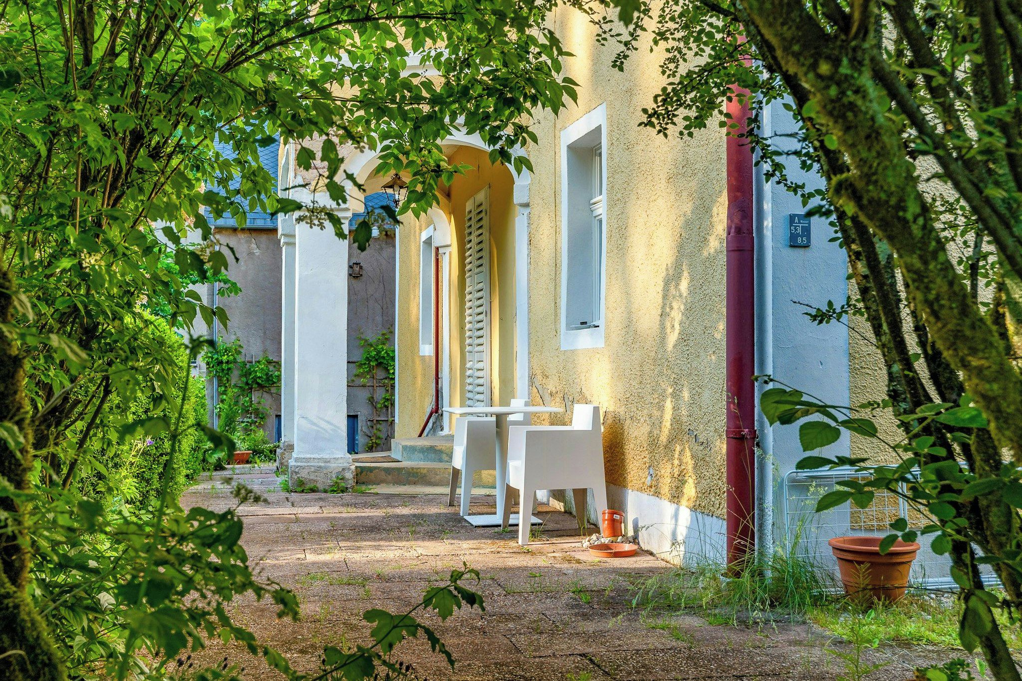 Une cour avec une table et des chaises blanches, un escalier et un bâtiment à la façade jaune.