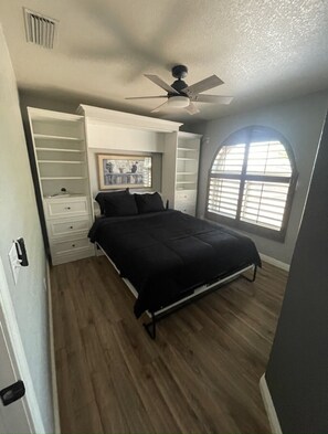 Ein Schlafzimmer mit einem Bett, einem Deckenventilator, einem Fenster mit Jalousien und integrierten Regalen.