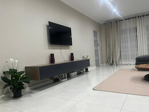 Ein modernes Wohnzimmer mit einem an der Wand befestigten Flachbildfernseher, einem dunklen Konsolentisch mit Dekoration, einer Topfpflanze und einem großen Fenster mit transparenten Vorhängen.