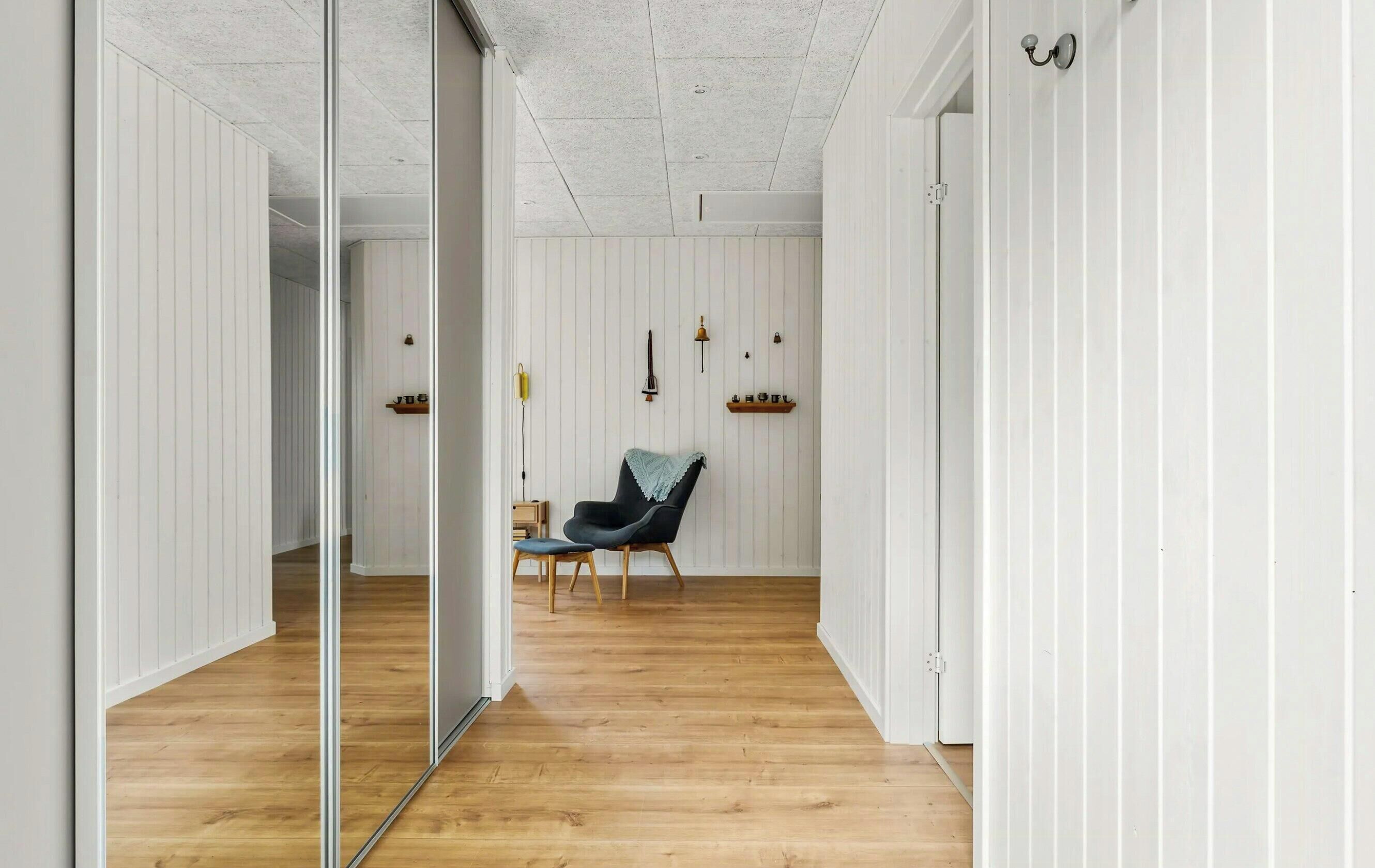 A hallway with wooden flooring, white paneled walls, a black chair, and a mirror.