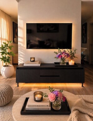 Ein modernes Wohnzimmer mit einem wandmontierten Flachbildfernseher, einer schwarzen Unterhaltungsanlage und einem runden Couchtisch mit Büchern und Kerzen.