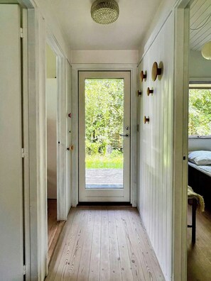 A hallway with a wooden floor, a door leading to a garden, and a bed with a white headboard in the background.