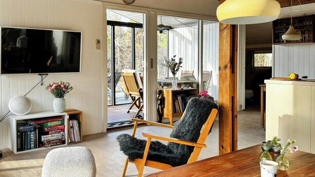 A modern living room with a wooden dining table, a fluffy chair, a sofa, and a TV mounted on the wall. - Charming family summerhouse near the beach (Højby)