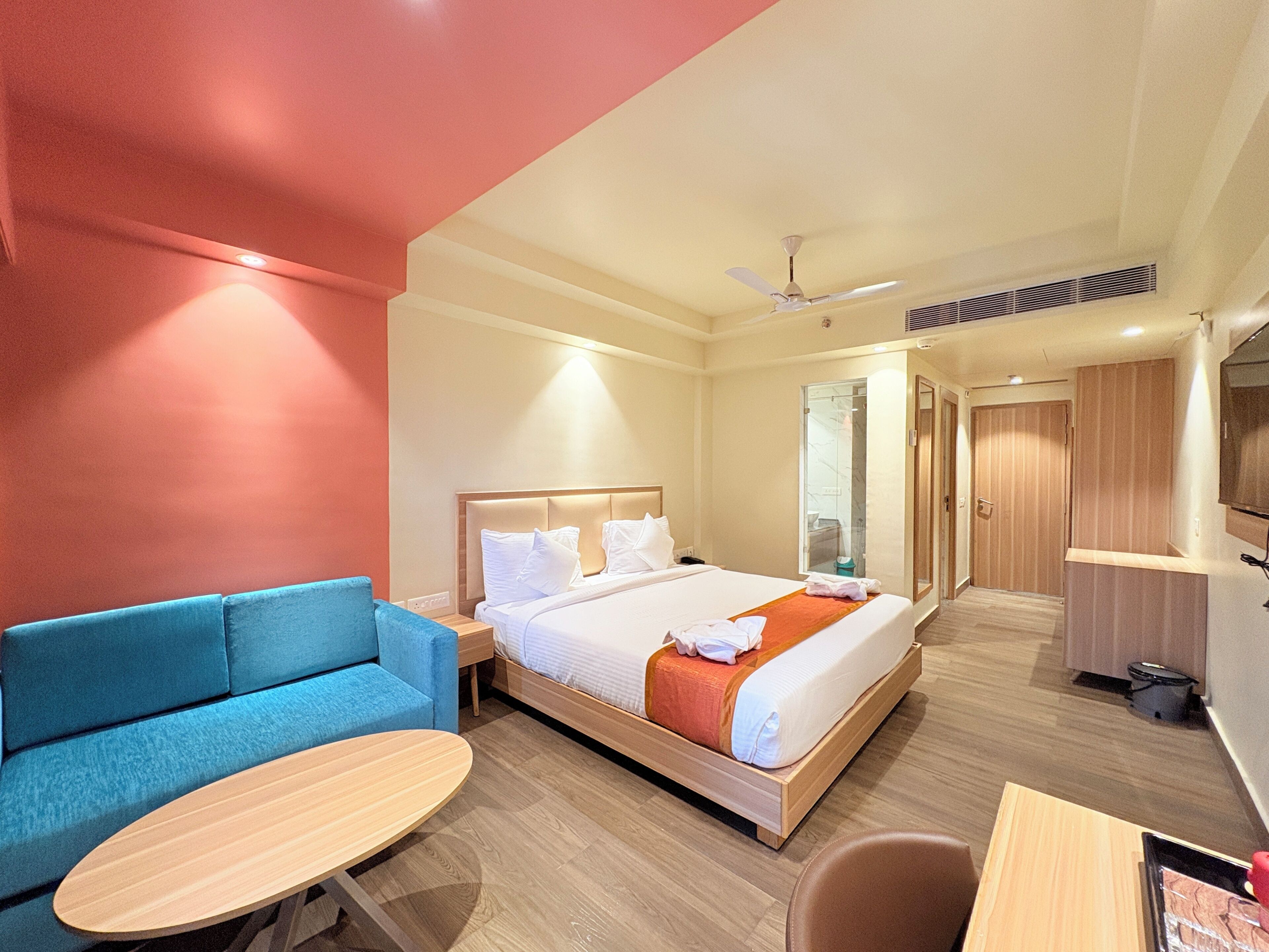 A hotel room with a bed, a blue sofa, a small table, a ceiling fan, and a TV.