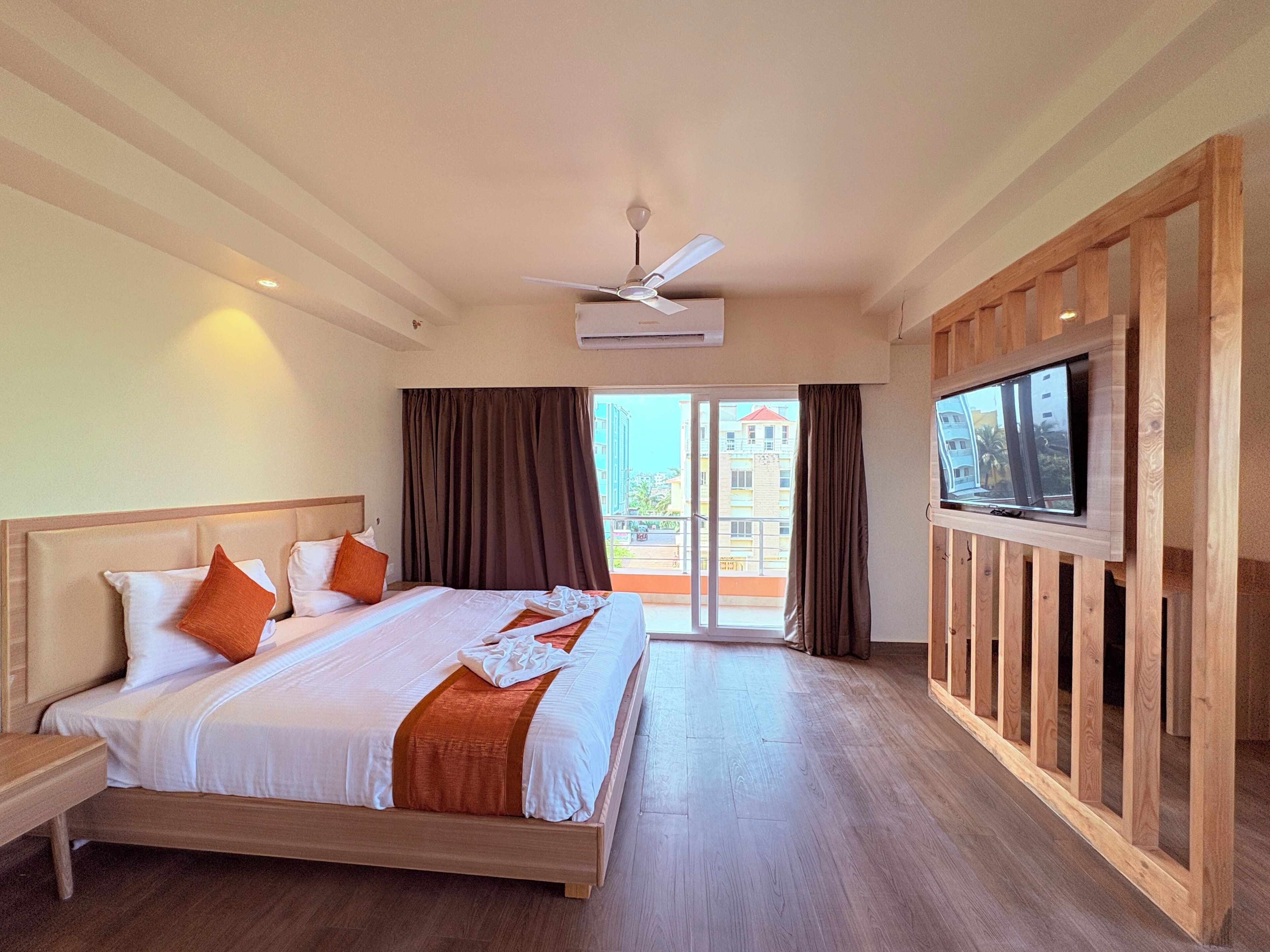 A hotel room with a bed, a ceiling fan, a TV, and a view of buildings outside.