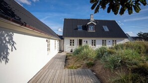 Una casa blanca con techo oscuro, una terraza de madera y un jardín con hierbas altas.