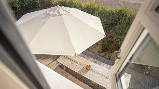 Una terraza de madera con una sombrilla blanca, una cesta de mimbre y un banco de madera.