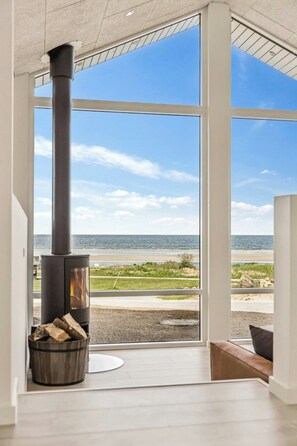 Un interior moderno con un ventanal grande que da a una playa, una chimenea con leña y un banco de madera.