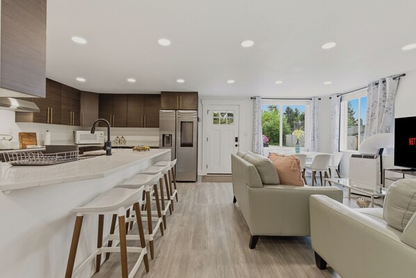 A modern kitchen with a central island, a dining area with bar stools, a living room with a sofa, and a TV. - Spacious 5BR Home Near Seattle & SeaTac - Parking (Burien)
