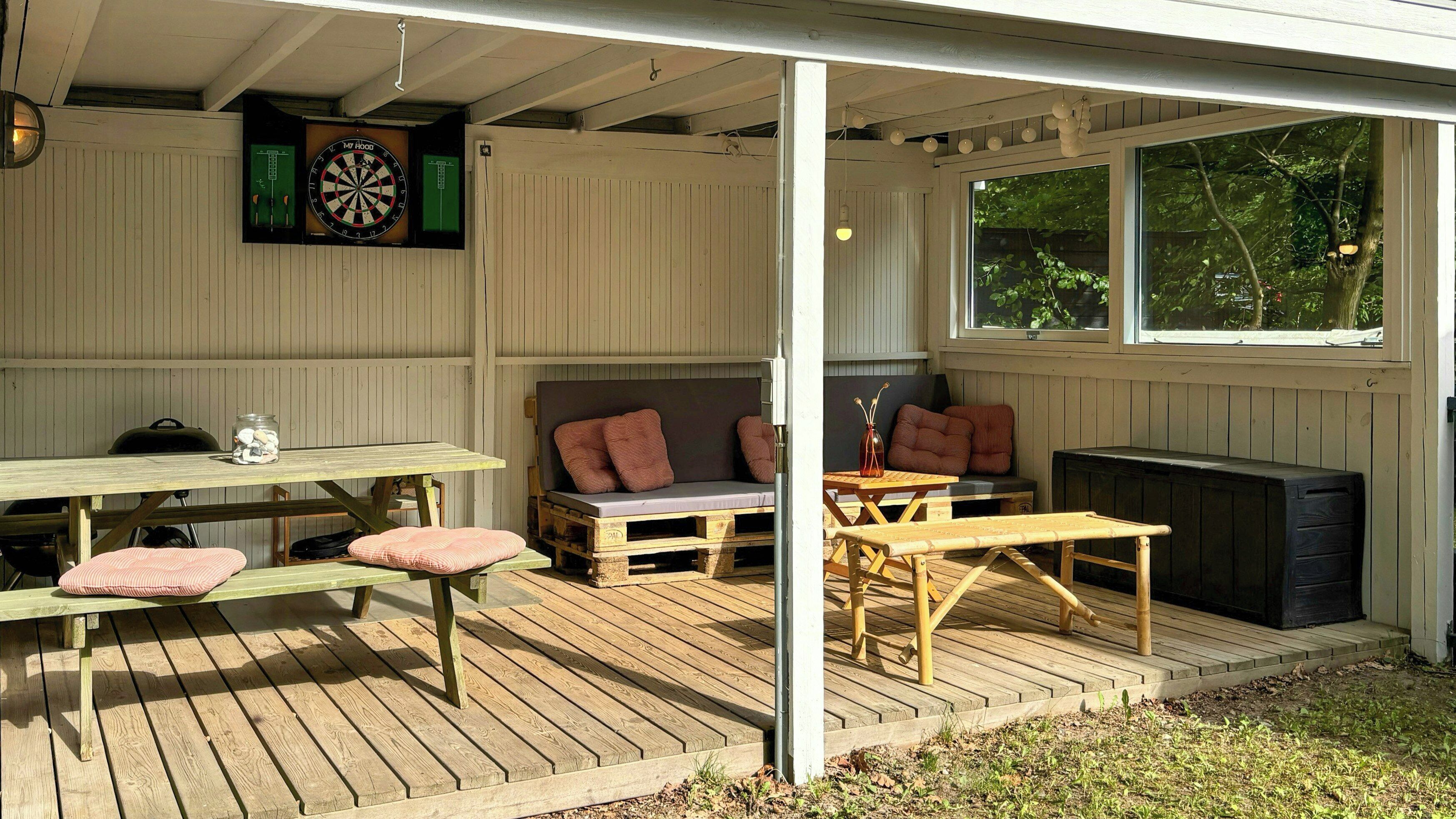 Et overdækket udendørsområde med træterrasse, et bord, en bænk, en sofa og en dartskive.