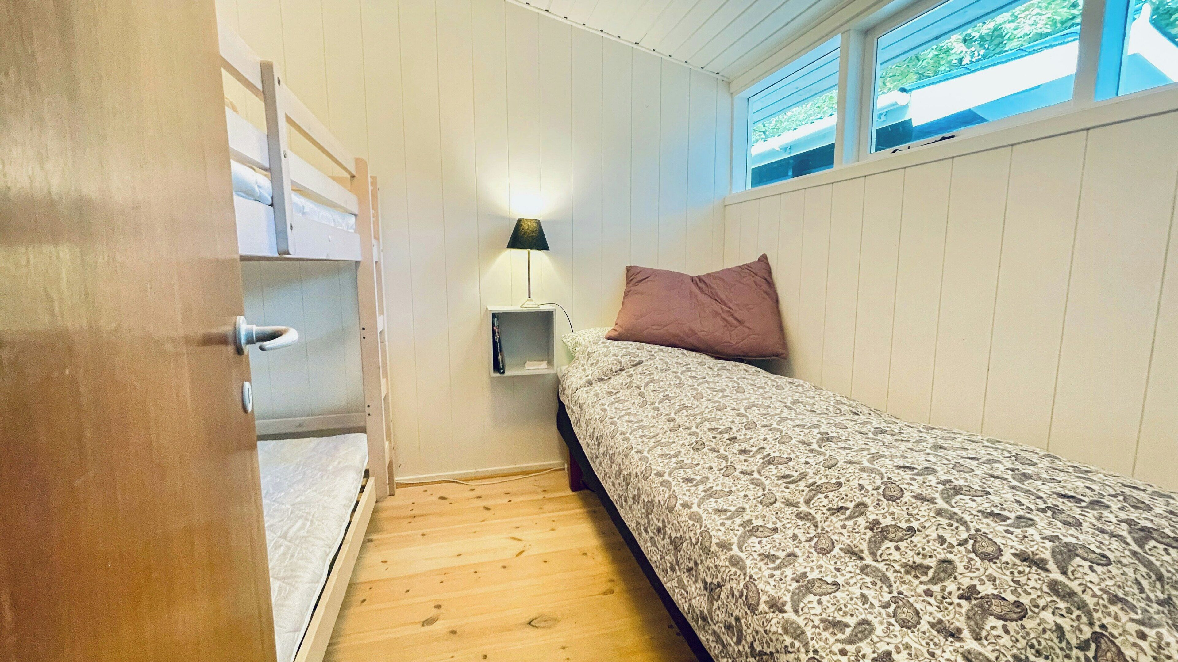 Ein kleines Schlafzimmer mit Holzboden, einem Bett mit gemusterten Betttextilien, einem Nachttisch mit Lampe und einem Fenster mit Blick auf Grünflächen.