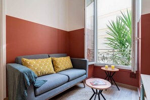 Ein Zimmer mit einer grauen Couch, einem kleinen runden Tisch mit Getränken und einem Fenster mit Blick auf Pflanzen.