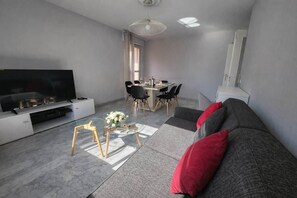 Ein modernes Wohnzimmer mit Flachbildfernseher, Essbereich mit Tisch und Stühlen, einer Couch mit roten Kissen und einem Glastisch.