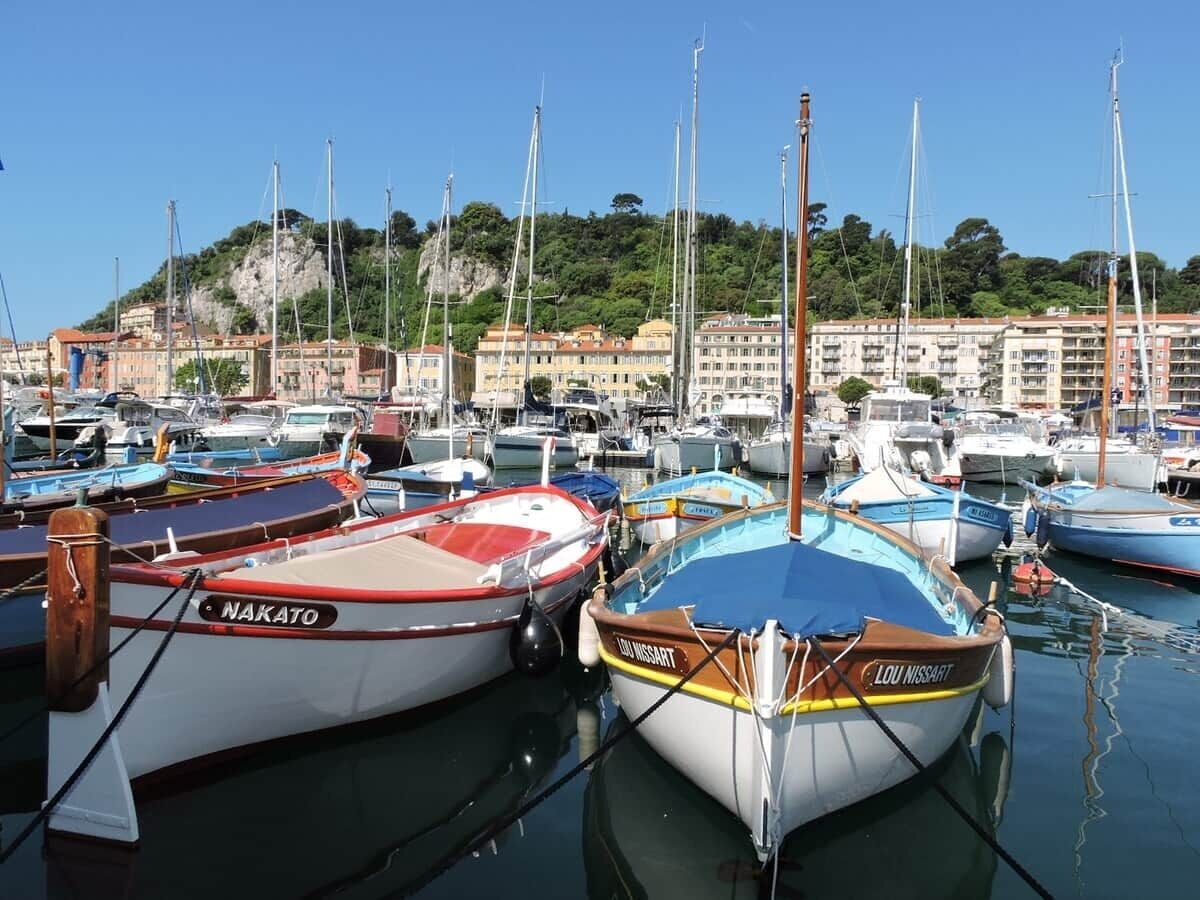 Ein Yachthafen mit vertäuten Booten, ein Gebäude am Wasser im Hintergrund und ein Hügel mit Grünpflanzen.