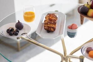 Ein Tisch mit einem Glas Orangensaft, einem Teller mit Feigen und einem Brotlaib sowie einer Obstschale.