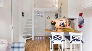 Uma cozinha compacta com piso de madeira, armários brancos, uma pequena mesa de jantar com duas cadeiras, uma escada e um porta-roupas fixado na parede.