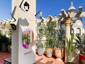 Ein Innenhof mit Blumentöpfen, eine weiße Säule mit rundem Spiegel und ein Gebäude mit Kuppel und dekorativen Säulen im Hintergrund.