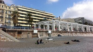 Una playa con una zona de estar cubierta, un muro de piedra y un edificio con balcones al fondo.