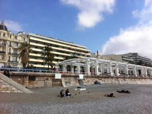 Ein Strand mit einer überdachten Sitzgelegenheit, einer Steinmauer und einem Gebäude mit Balkonen im Hintergrund.