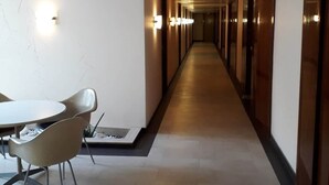 Un pasillo largo y estrecho con una mesa y sillas a la izquierda, lámparas en las paredes y suelo de baldosas.