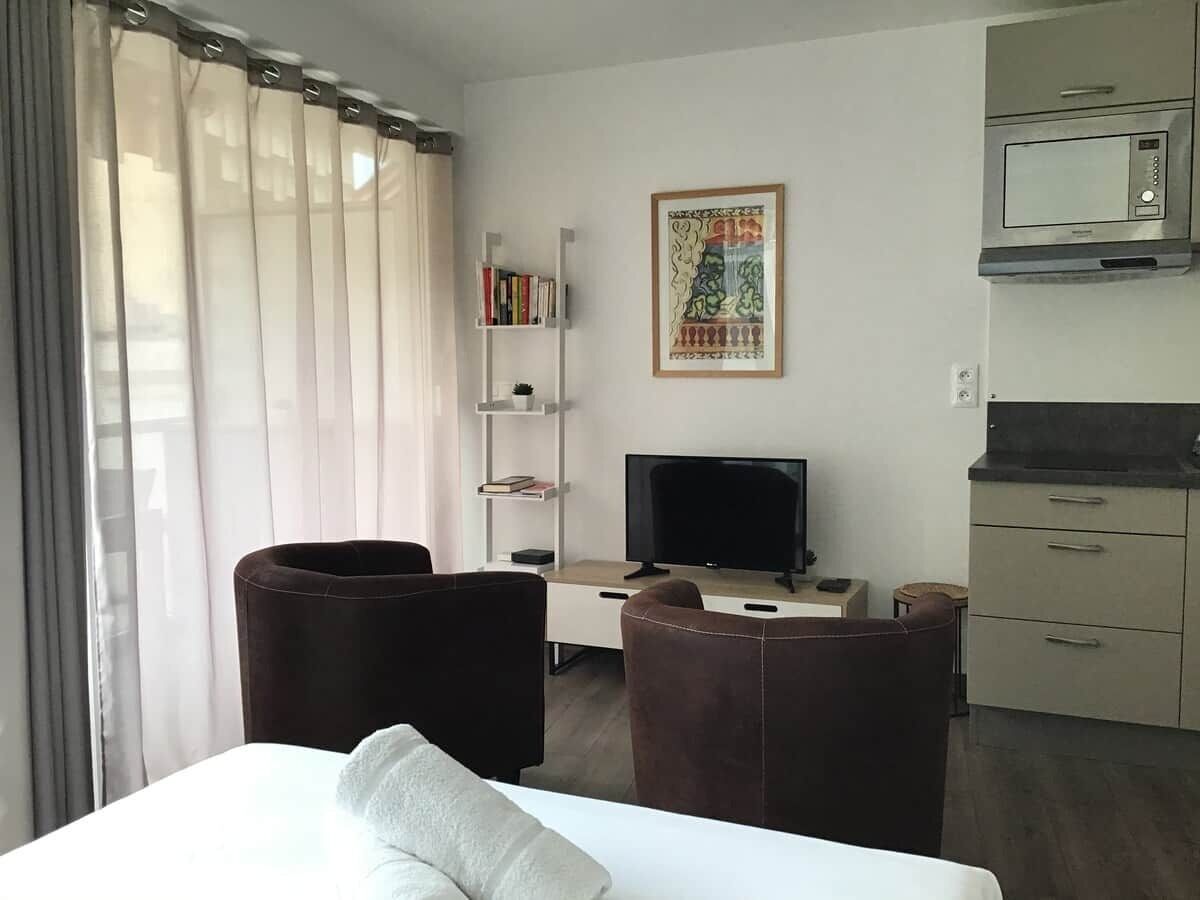 A hotel room with a bed, two chairs, a desk with a TV, a bookshelf, a painting, and a kitchenette.