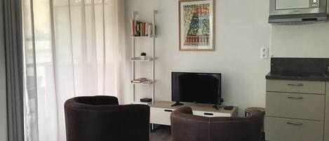 A hotel room with a bed, two chairs, a desk with a TV, a bookshelf, a painting, and a kitchenette.