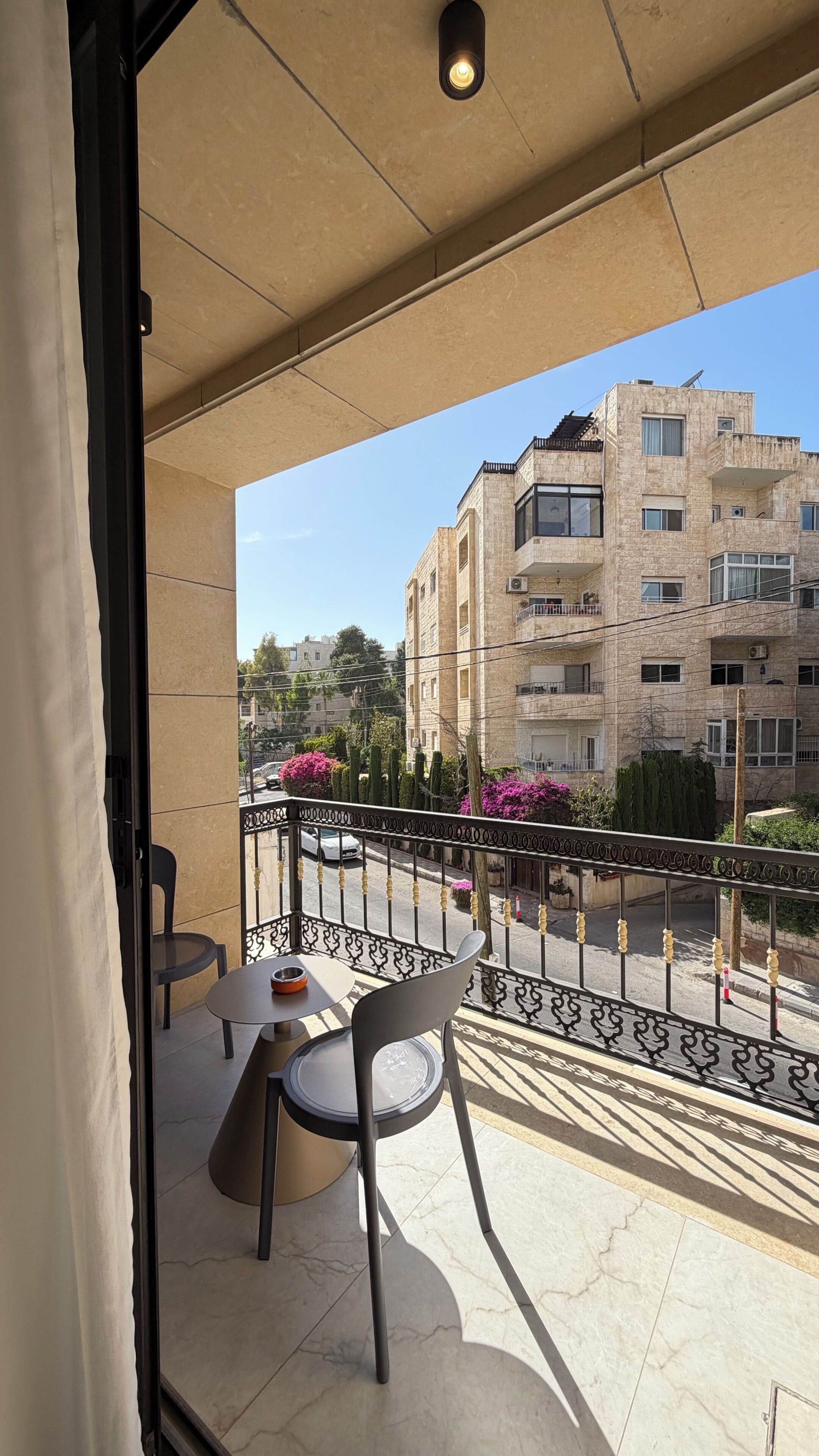A balcony with a table and chairs, a view of a building, and a street with cars and plants.
