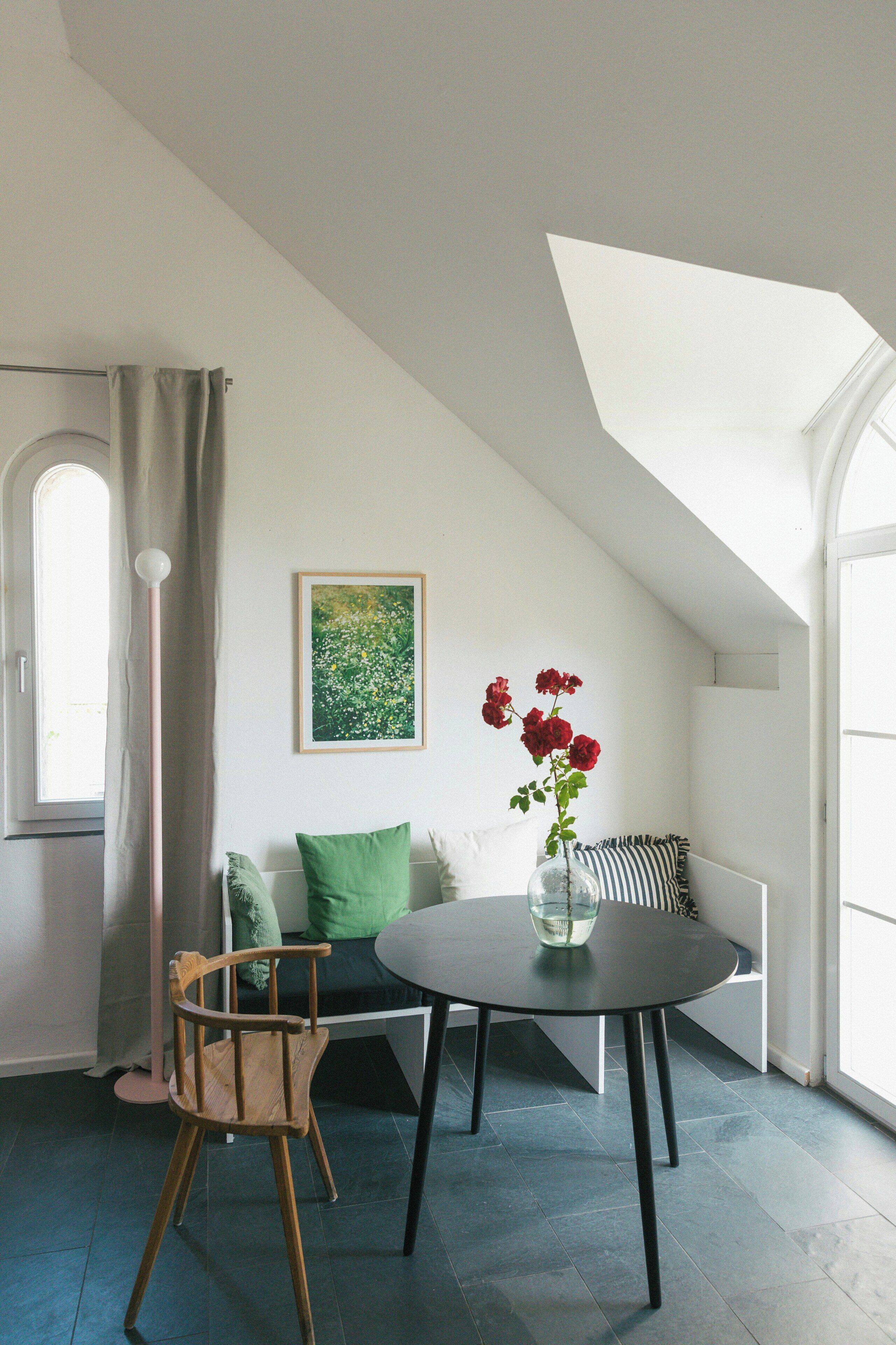 A room with a round table, a wooden chair, a sofa, a green pillow, a vase with flowers, a framed picture, and a window with curtains.