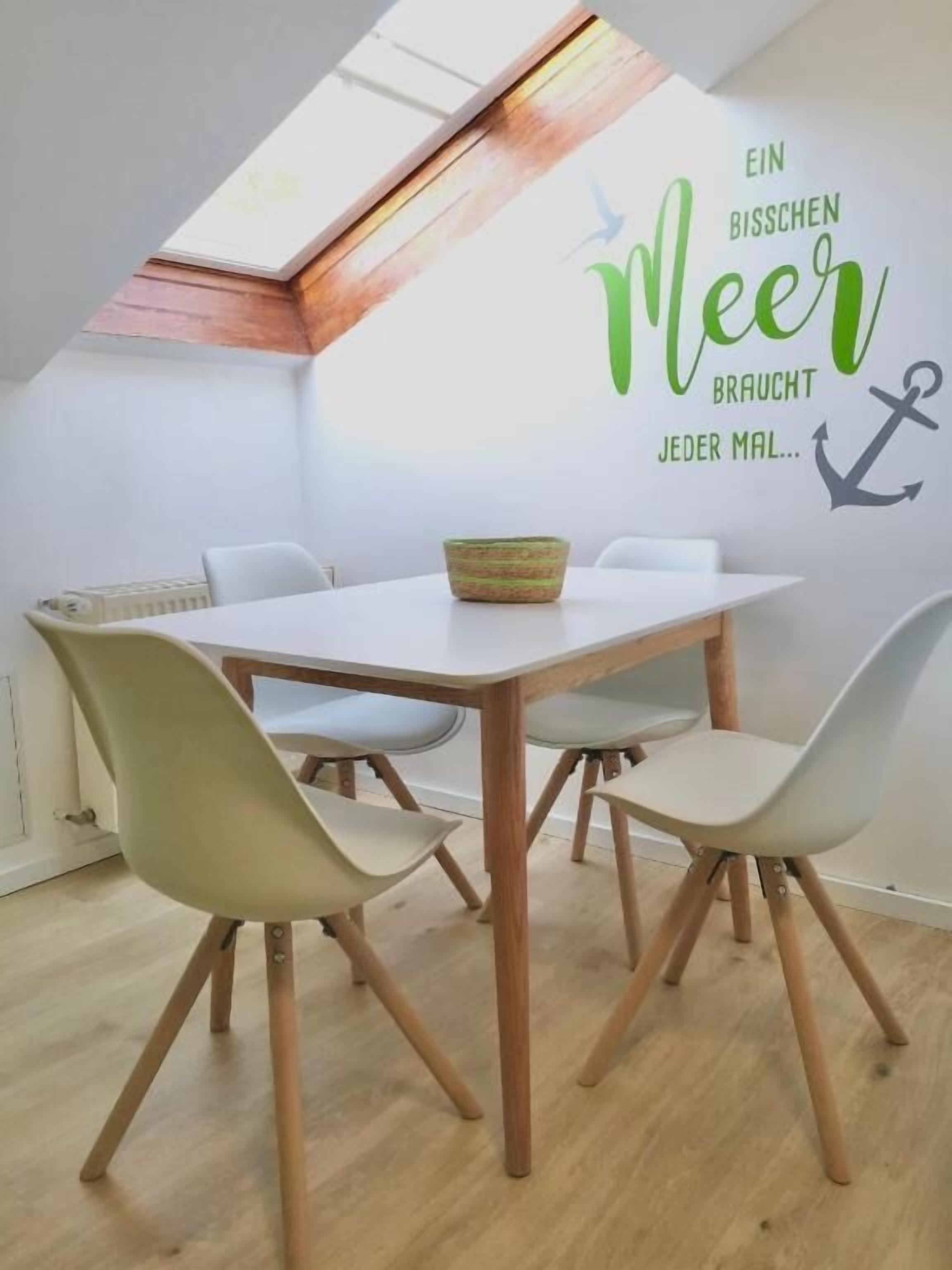 A dining area with a white table, four chairs, and a wall decal with text and an anchor.